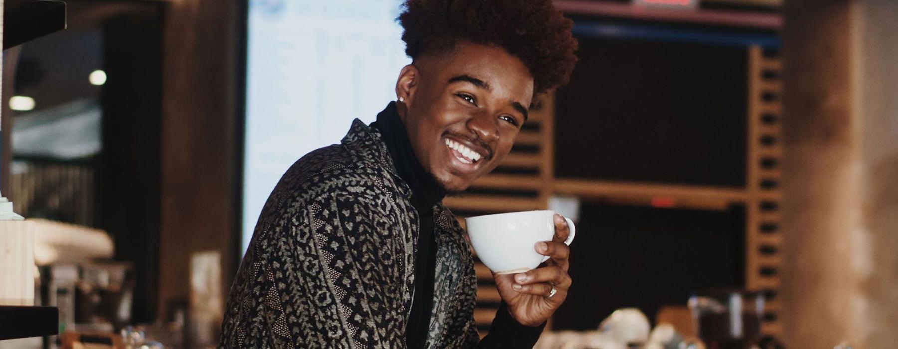 young man smiles while holding a cup of coffee in coffee shop